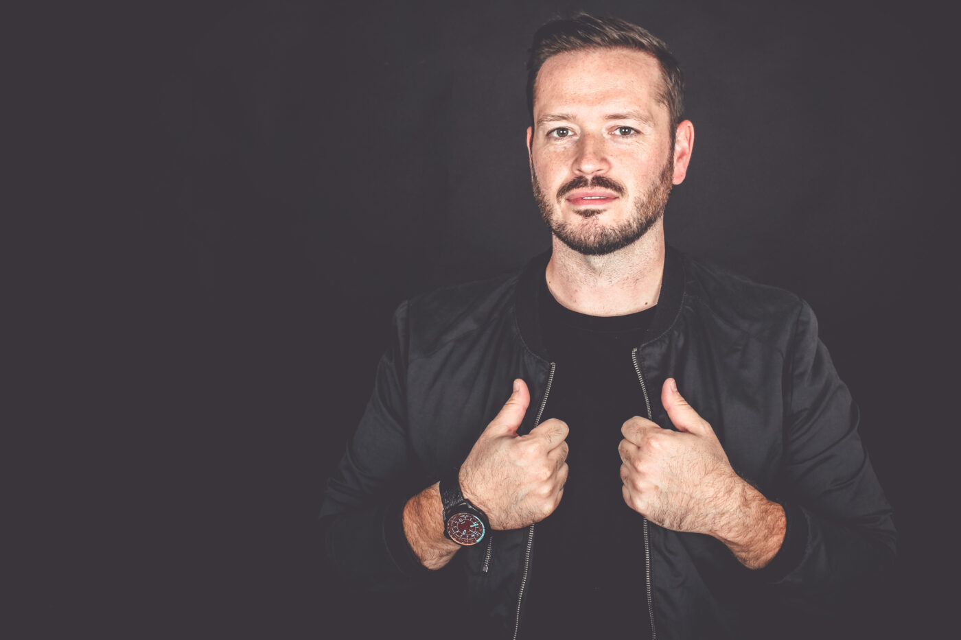 Benjamin Schäfer blickt präsent in die Kamera, die Hände halten die Revers seiner dunklen Jacke vor schwarzem Hintergrund.